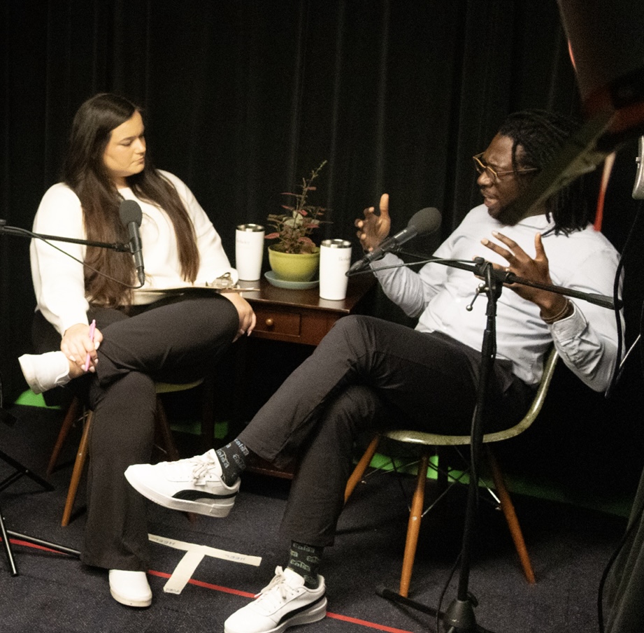 An African American man and student with long brown hair speak animately in a dark studio with microphones in front of them.